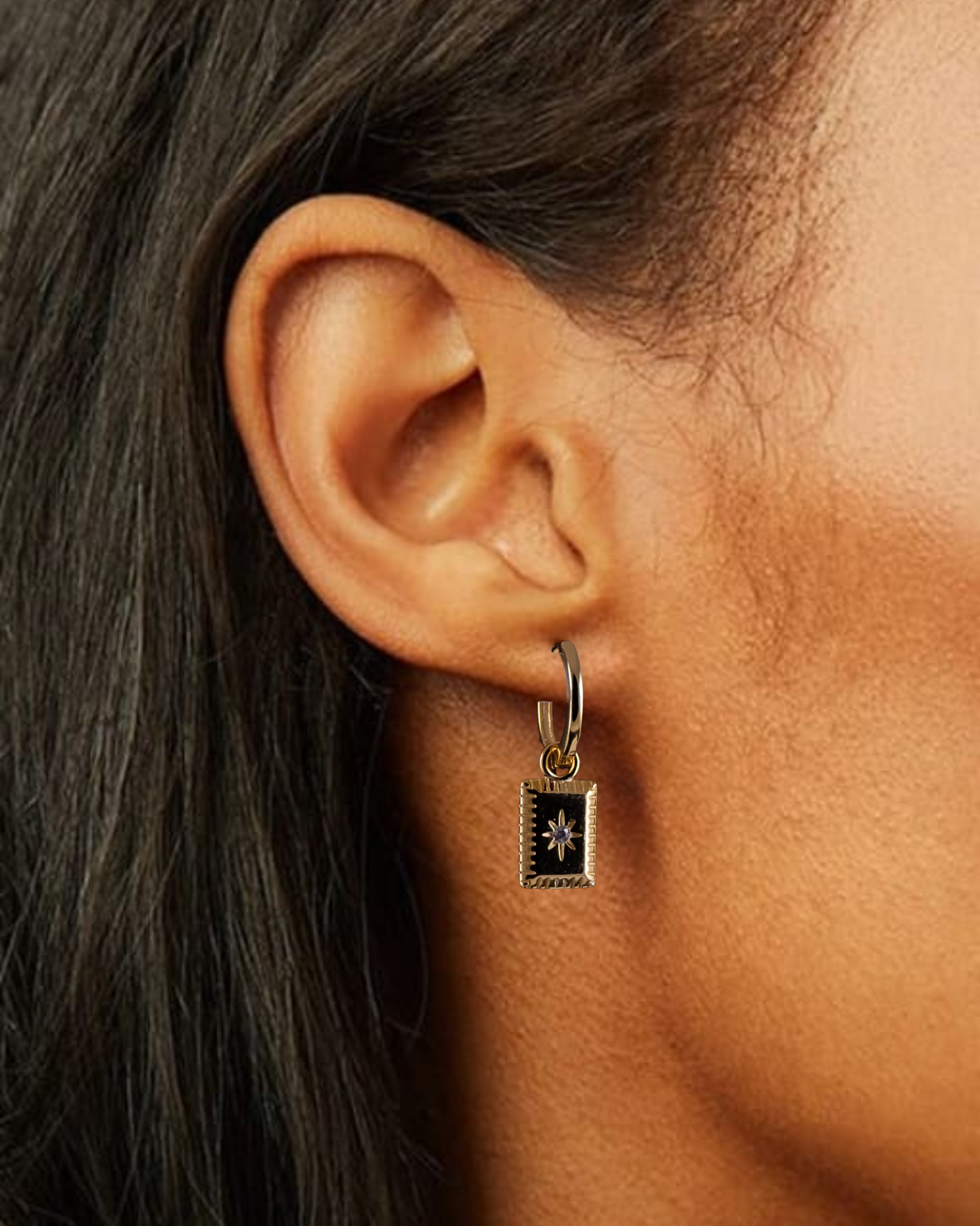 Close-up of an ear wearing a small hoop earring with a square pendant.