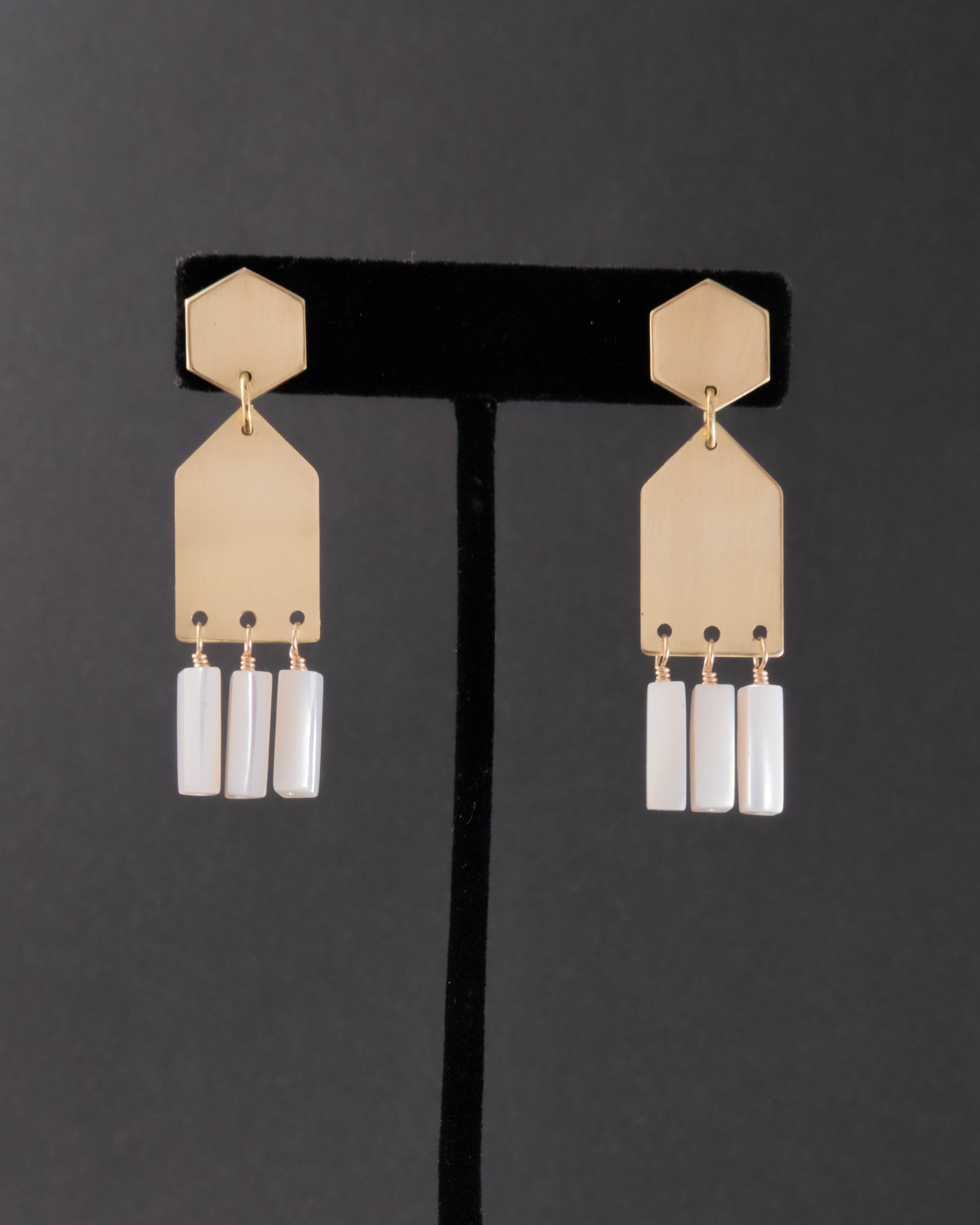 Gold earrings with mother of pearl gems on a black stand against a dark background