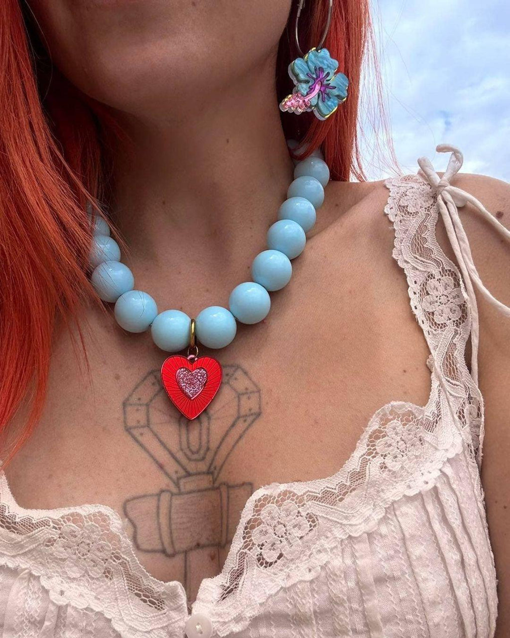 Person wearing a light blue beaded necklace with a red heart charm, floral earrings, and a white lace top against a blue sky.