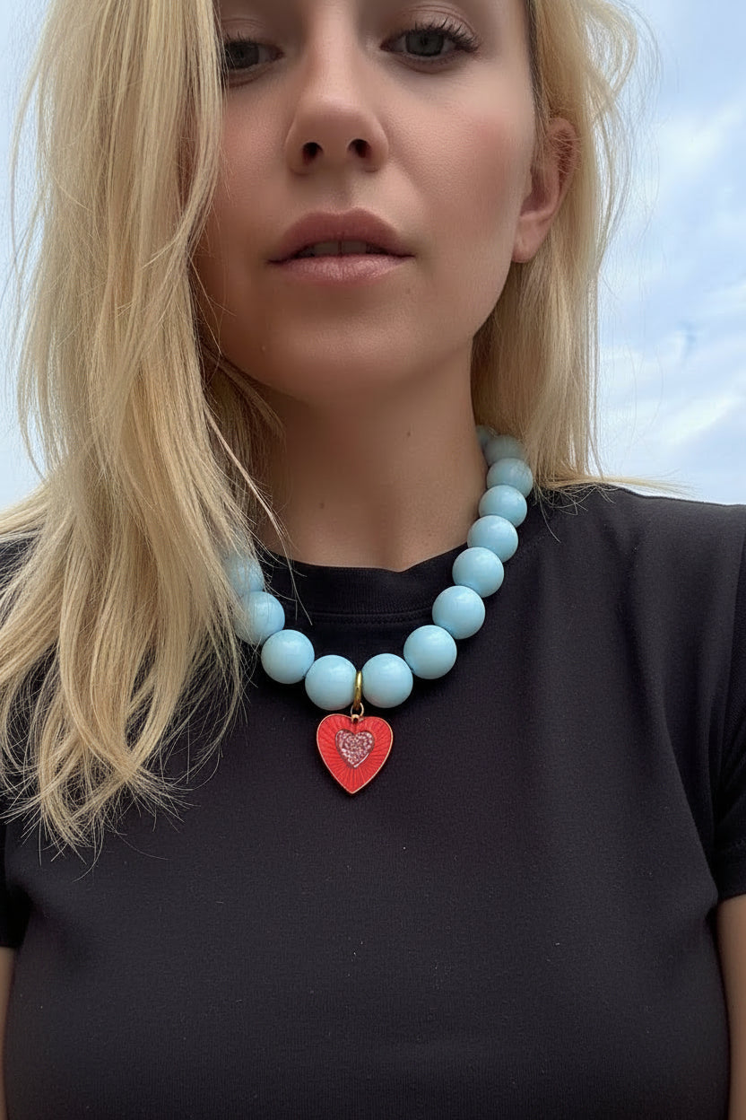 Person wearing a light blue beaded necklace with a red heart charm, floral earrings, and a white lace top against a blue sky.