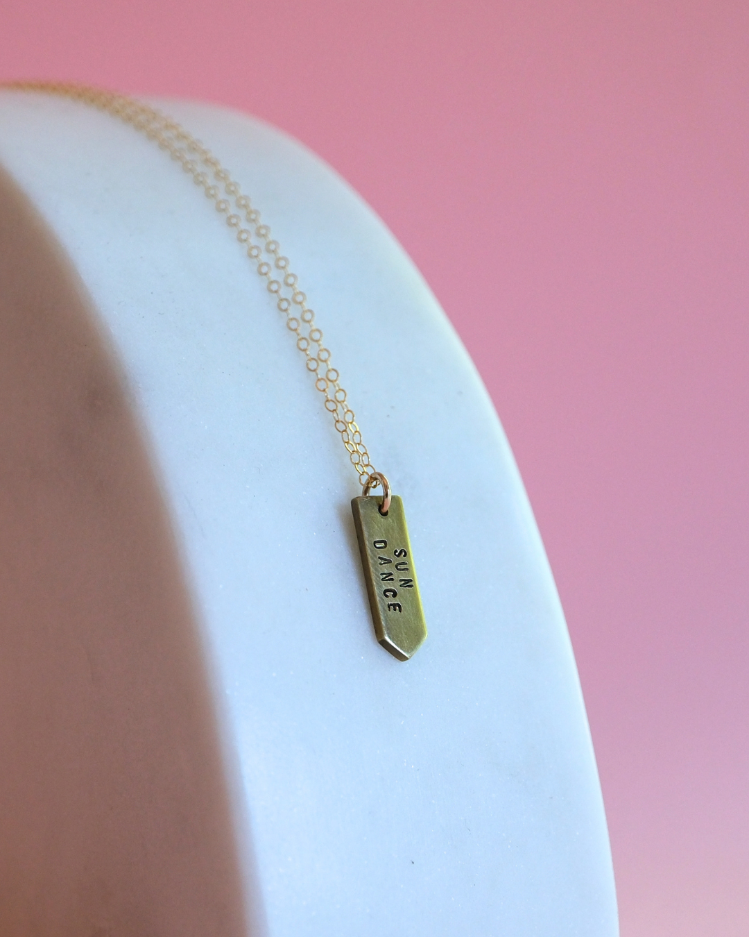 Gold necklace with a stamped pendant saying SUN DANCE on a white plate against a pink background
