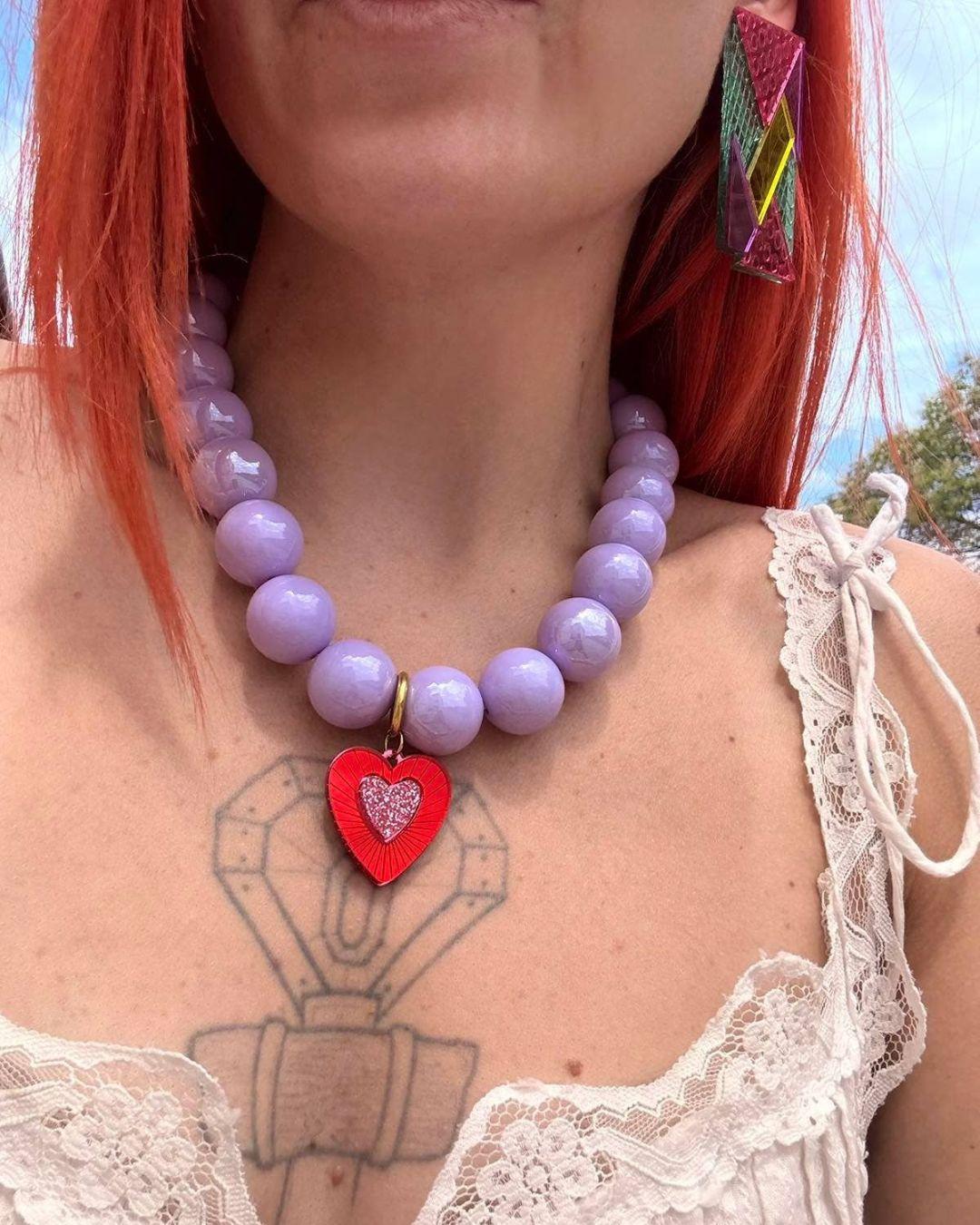 Person wearing a lilac beaded necklace with a red heart charm, outdoors.