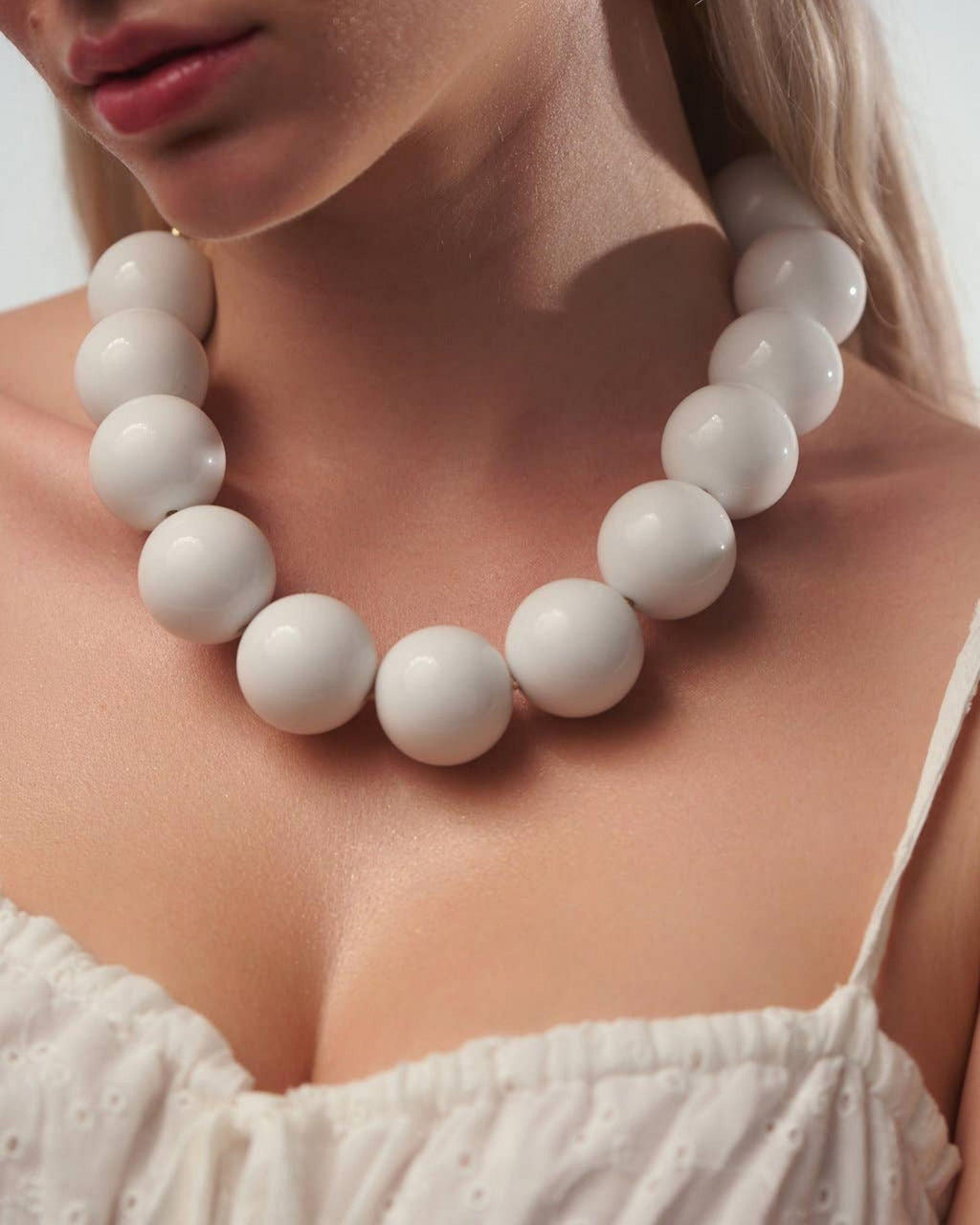 A close-up of a model wearing a chunky white beaded necklace with large, smooth spherical beads. The model is wearing a white eyelet top, and the background is a clean, neutral studio setting.