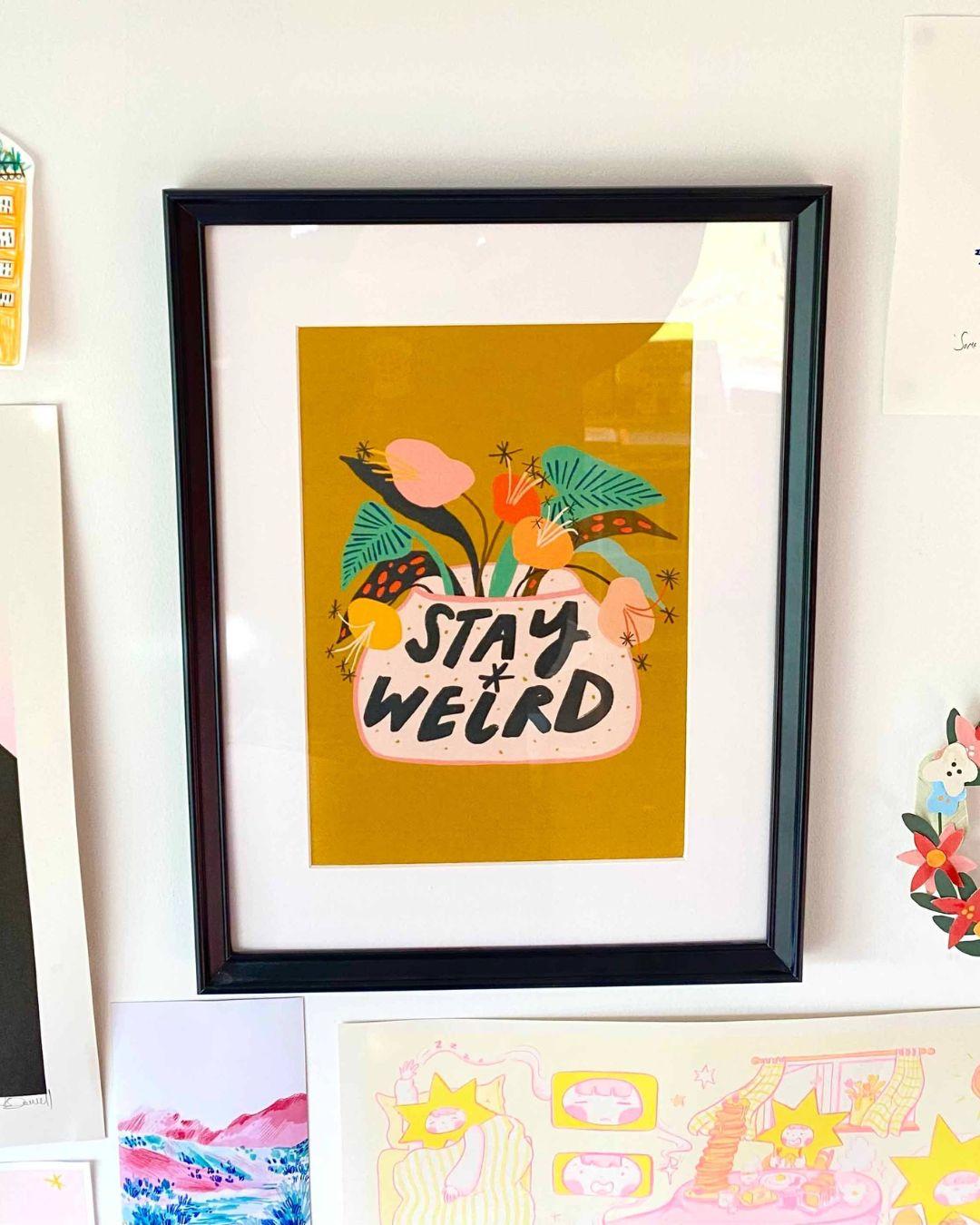 Framed artwork with 'Stay Weird' text and colorful plants on a yellow background