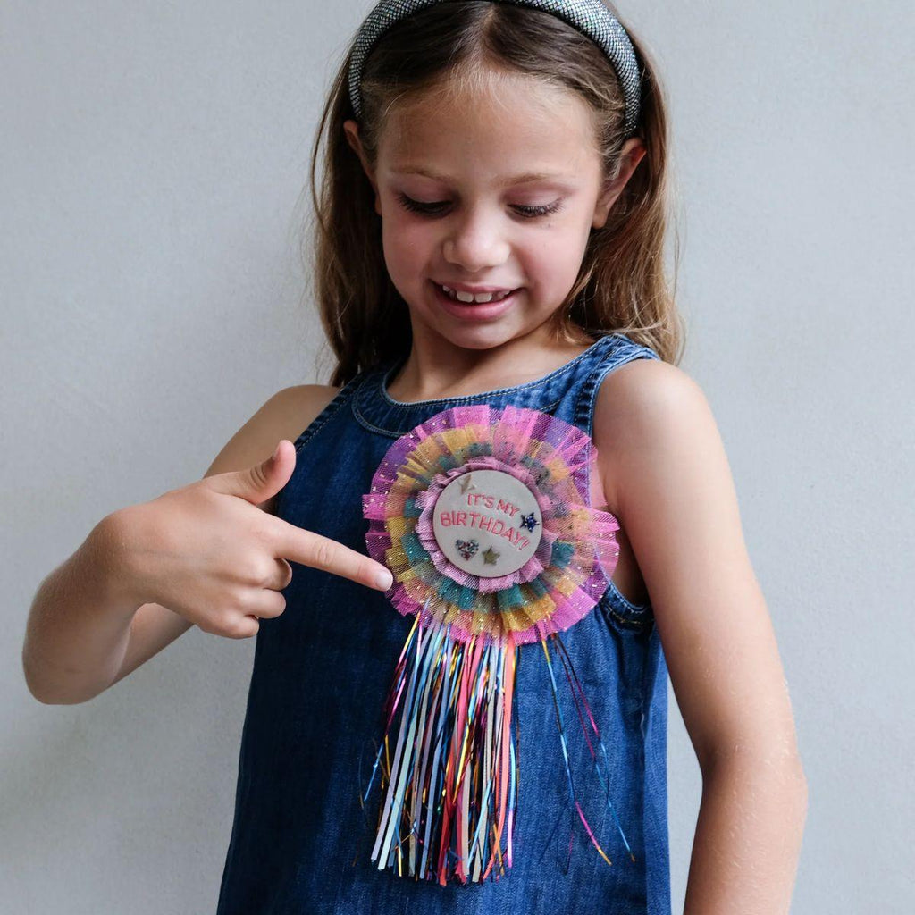 Young girl wearing a colorful birthday corsage and pointing at it against a plain background