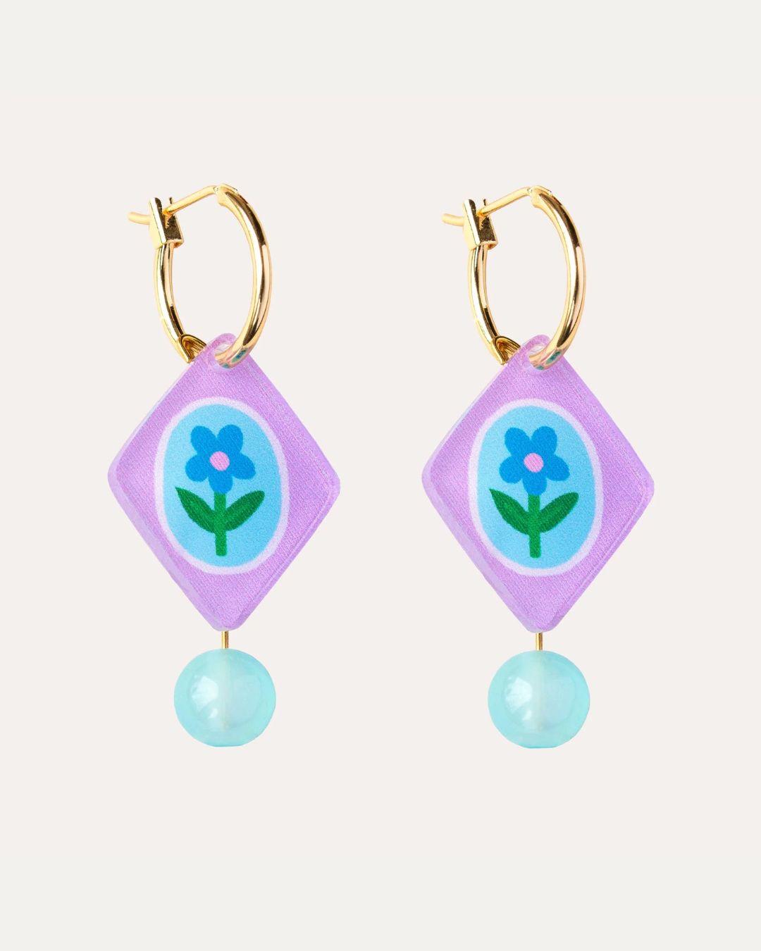 Pair of hoop earrings with purple diamond-shaped charms featuring a blue flower design on a white background.