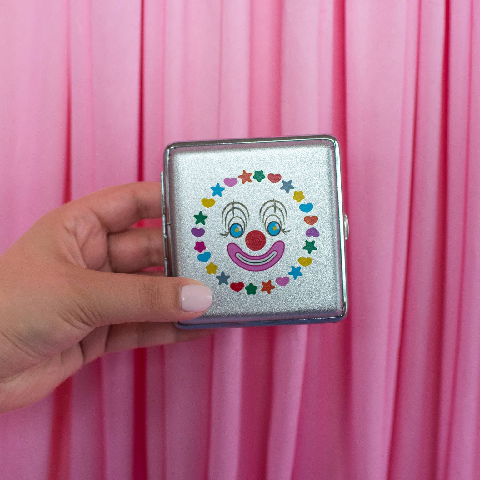 Hand holding a small rectangular object with a clown face design against a pink curtain background
