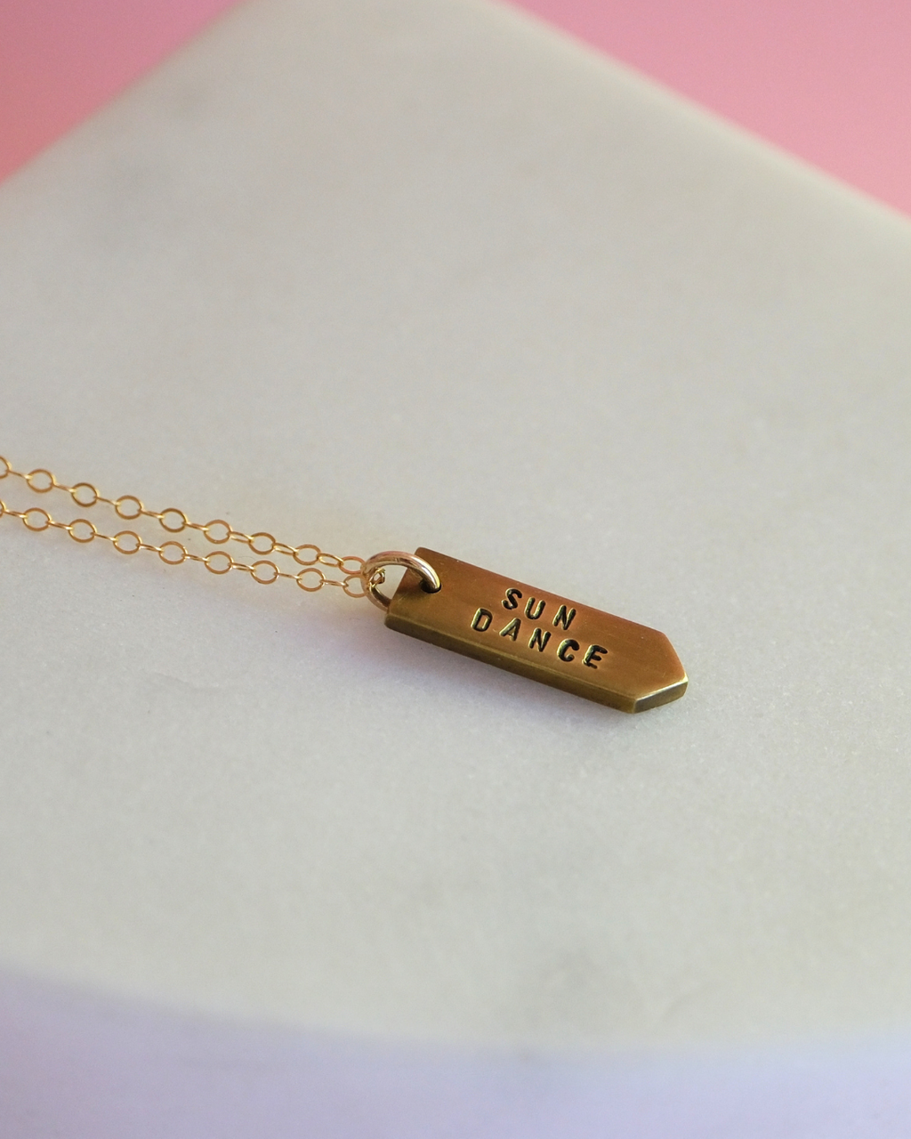 Gold necklace with 'SUN DANCE' pendant on a light pink background