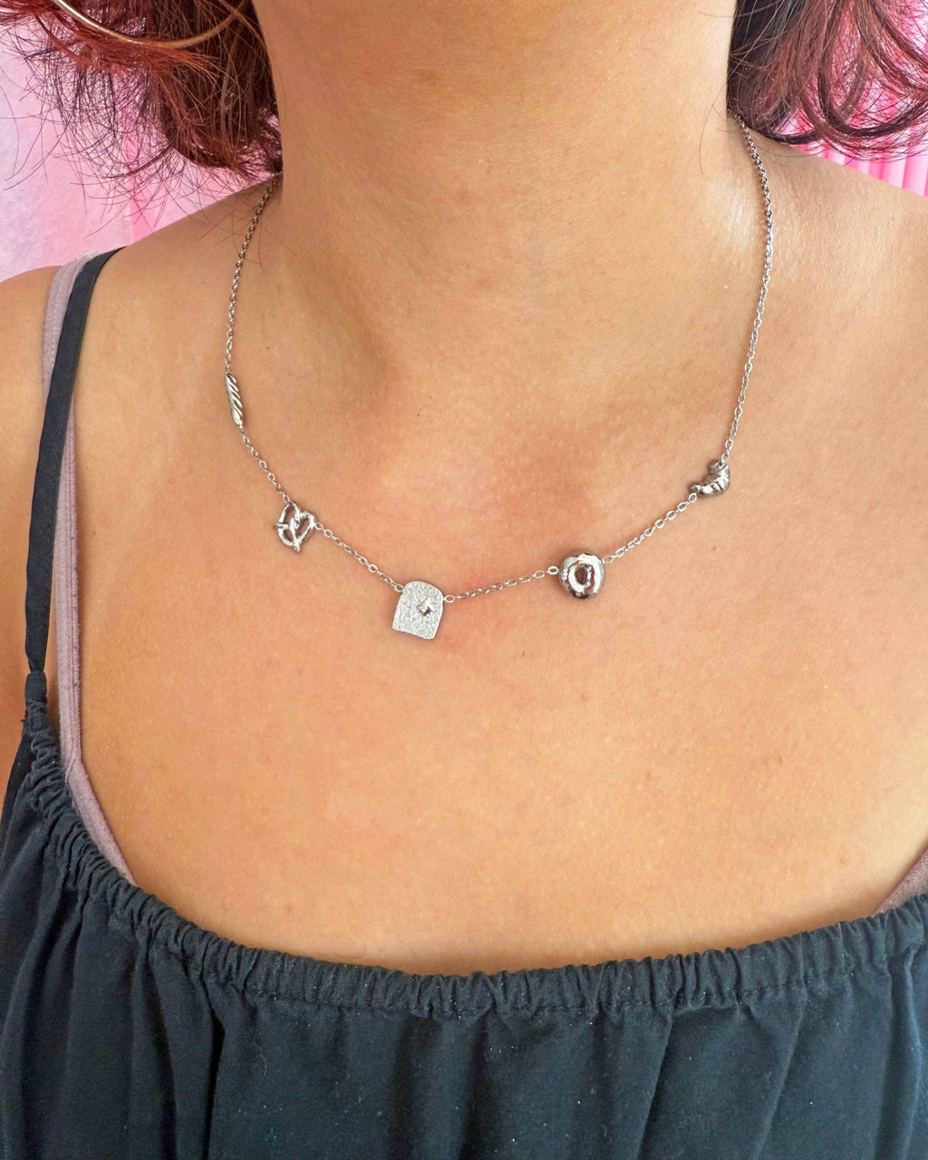 Silver necklace with small pendants on a person wearing a black top against a pink background