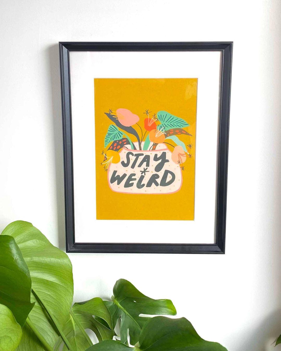 Framed artwork with 'Stay Weird' text and floral design on a yellow background, mounted on a white wall with green leaves in the foreground.