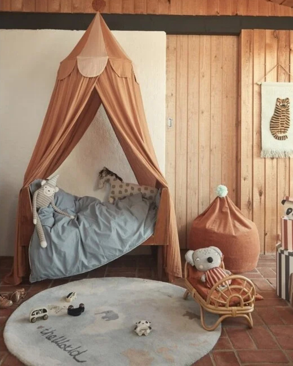 A cozy children's bedroom scene featuring a tall tan fabric canopy bed with a light blue duvet, a matching tan bean bag chair with a teddy bear, and a round gray rug on a tiled floor against a wood-paneled wall.