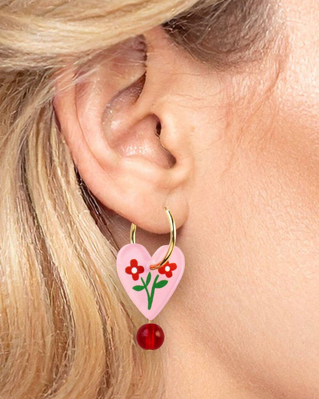 Heart-shaped earring with floral design and red bead worn by a person with blonde hair.