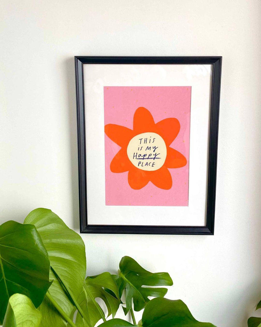 Framed artwork with a red flower and 'This is my happy place' text on a pink background, hanging on a wall with green leaves in the foreground.