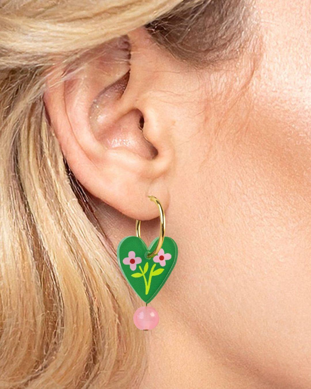 Heart-shaped earring with floral design and pink bead worn by a person with blonde hair.