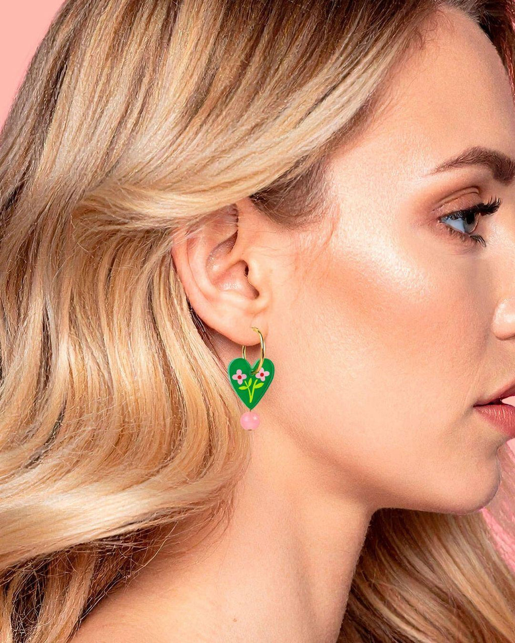 Close-up of a person wearing a green heart-shaped earring with floral details against a pink background