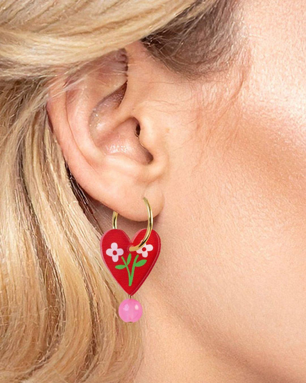 Heart-shaped earrings with floral design and pink bead worn by a person with blonde hair.