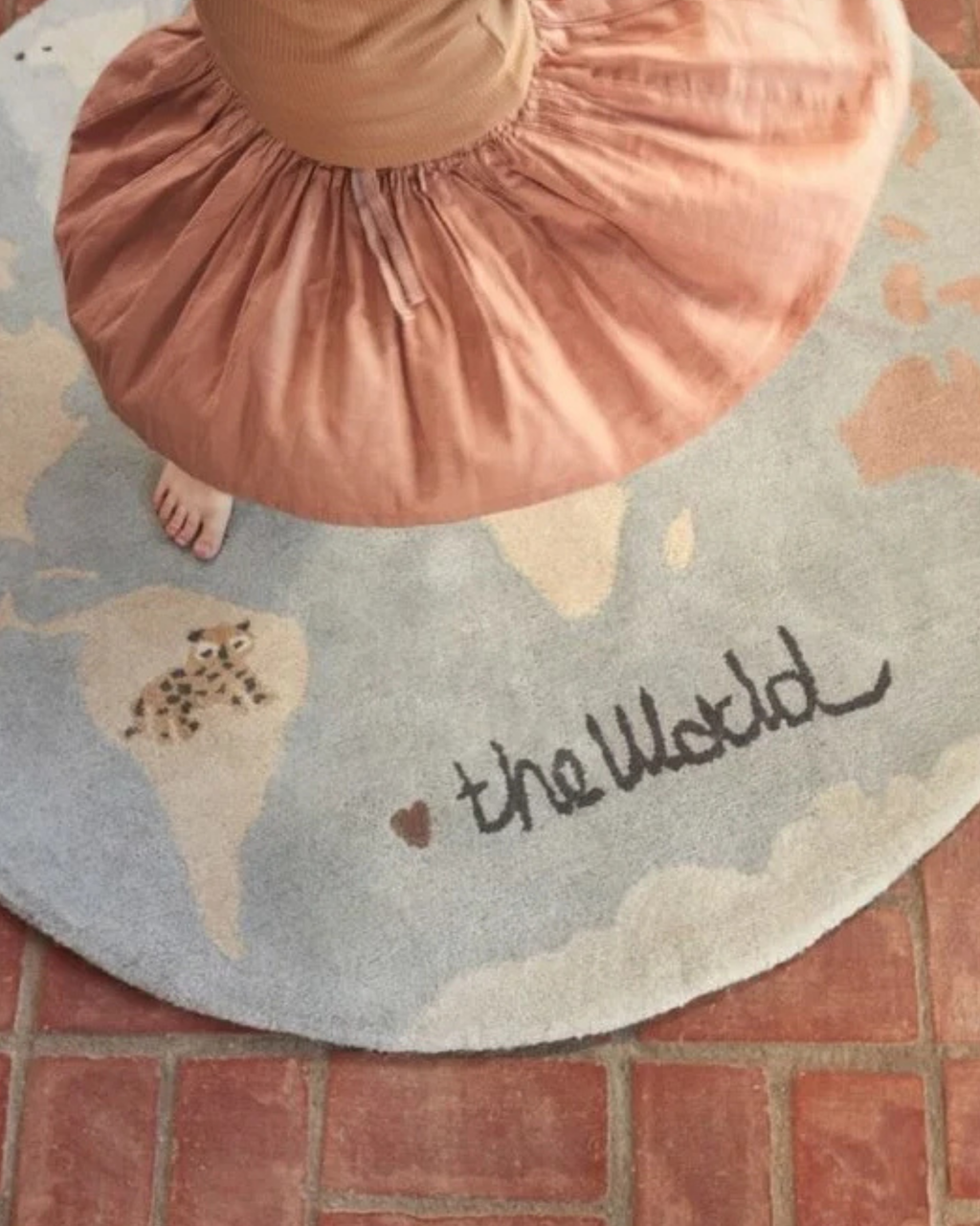 A round area rug with a world map design in light blue and beige, featuring the words 'the World' and a small heart. A person wearing a dusty pink skirt is standing on the rug, with their foot visible.