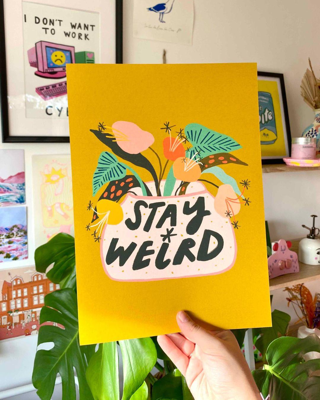 Hand holding a yellow card with 'Stay Weird' design against a wall with framed pictures and plants.