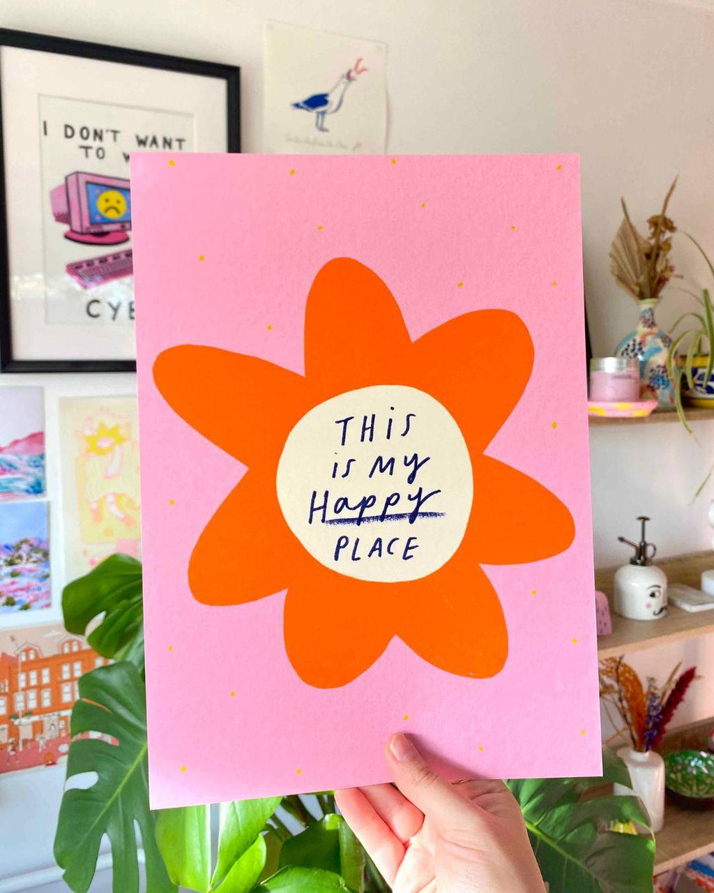Hand holding an art print with an orange flower and 'This is my happy place' text on a pink background.