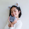 Young female presenting child wearing cat ears and holding a cat-shaped plush toy against a plain background