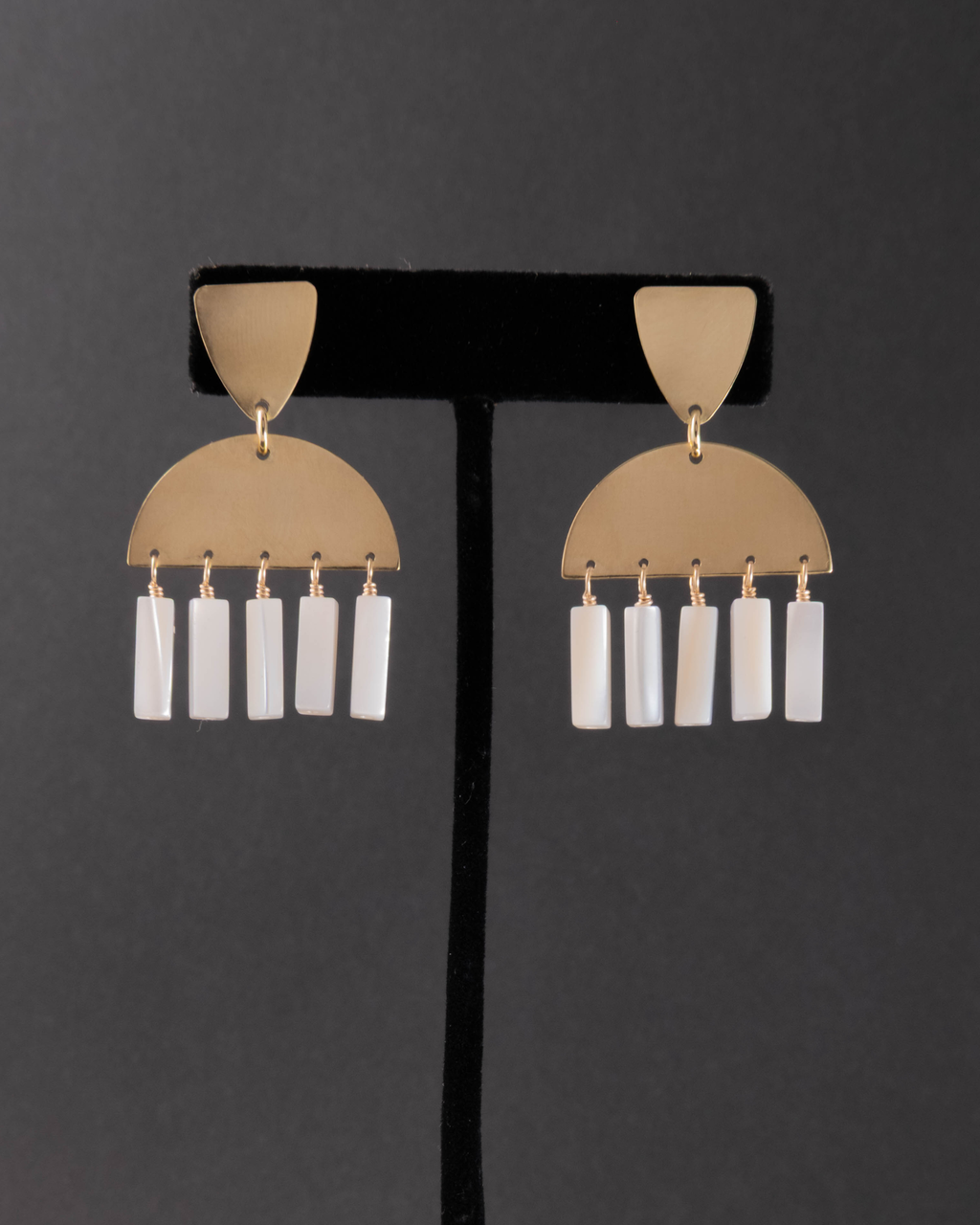 Pair of gold and mother of pearl earrings on a black stand against a dark background