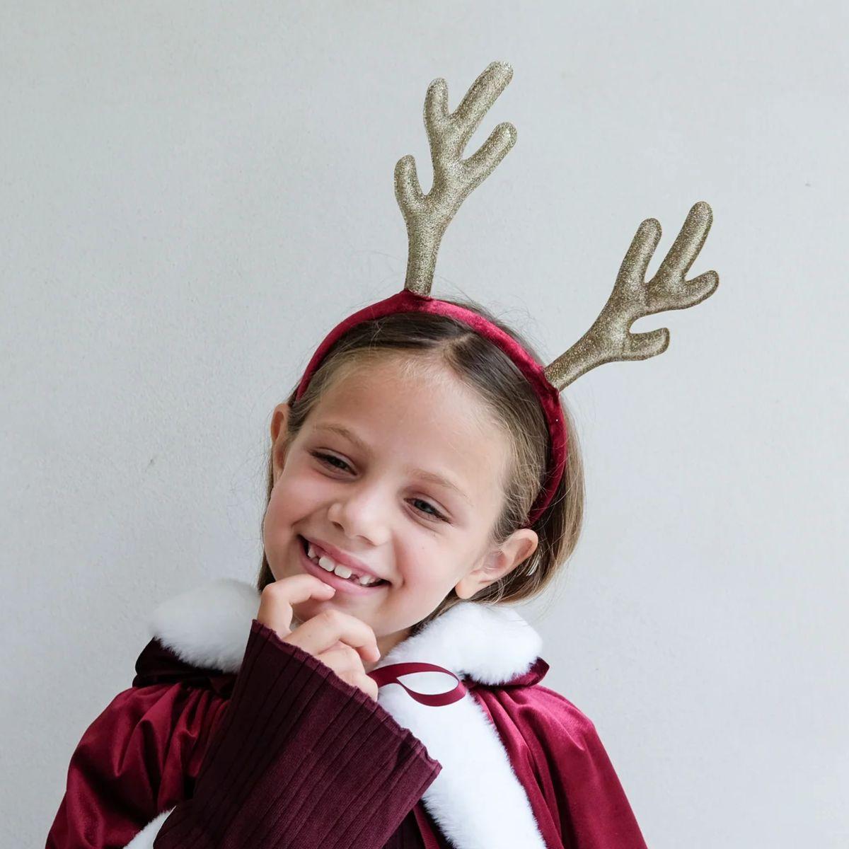 Child wearing a red coat and gold reindeer antlers against a plain background