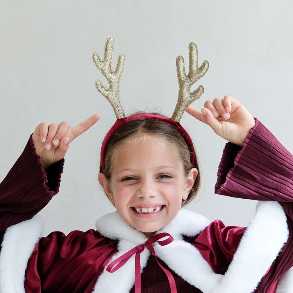 Child wearing a festive outfit with reindeer antlers and a white fur collar against a plain background