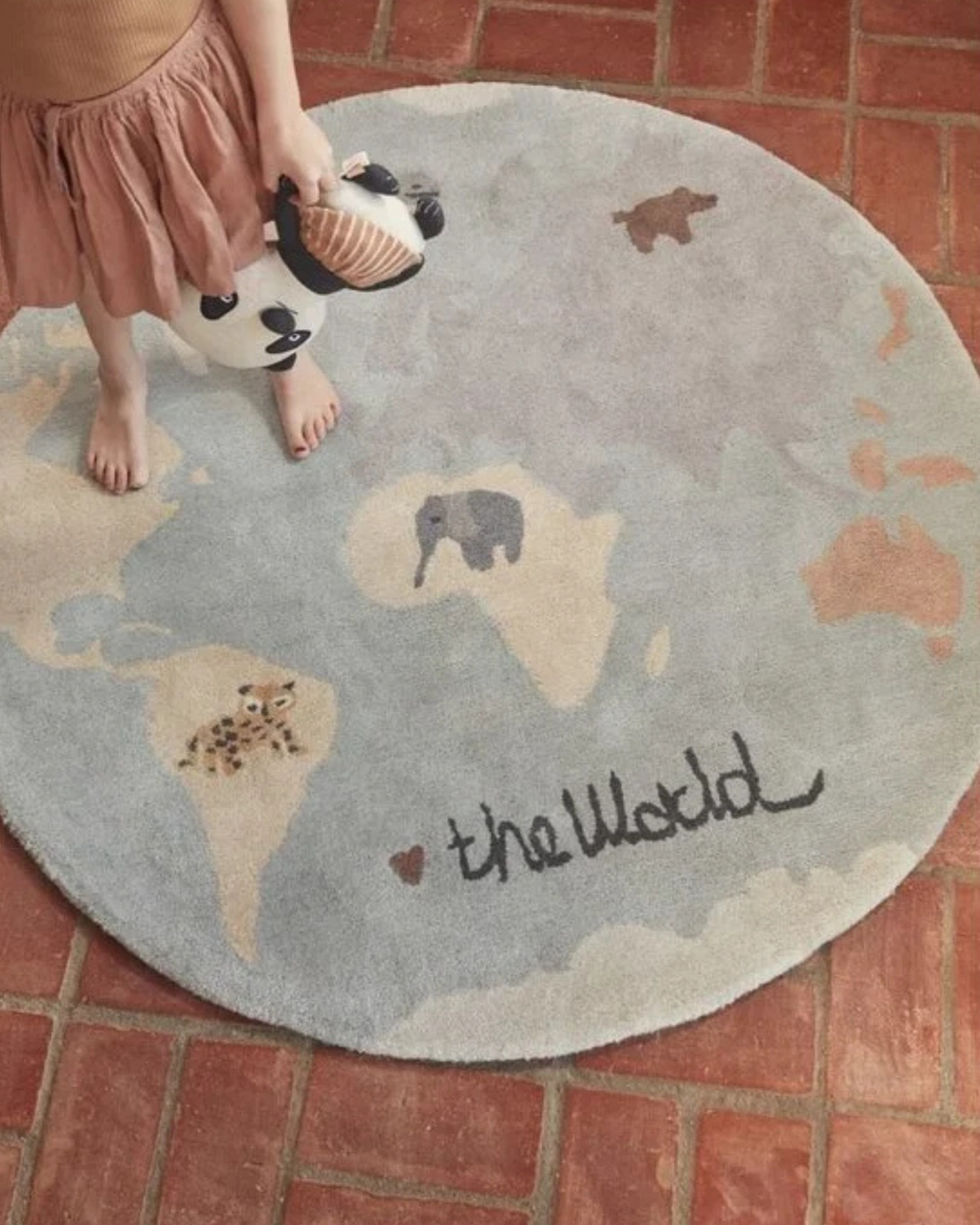 A round area rug with a world map design in muted blue-gray, cream, and tan, with the words 'the World' written in black script. A child's bare feet and skirt are visible standing on the rug, which is placed on a red brick floor.