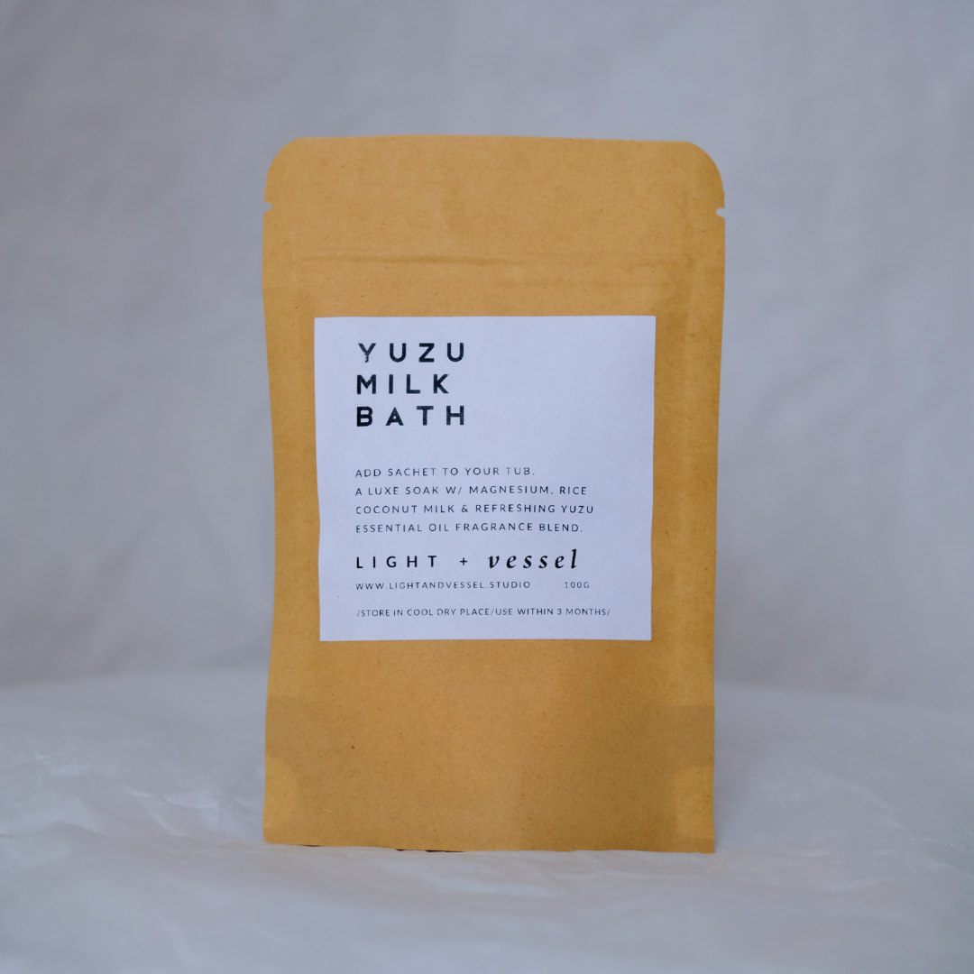 A package of Yuzu Milk Bath by Light + Vessel, with a white label on a yellow background, indicating the product is a bath additive with magnesium, rice coconut milk and yuzu essential oil, and 100g weight.