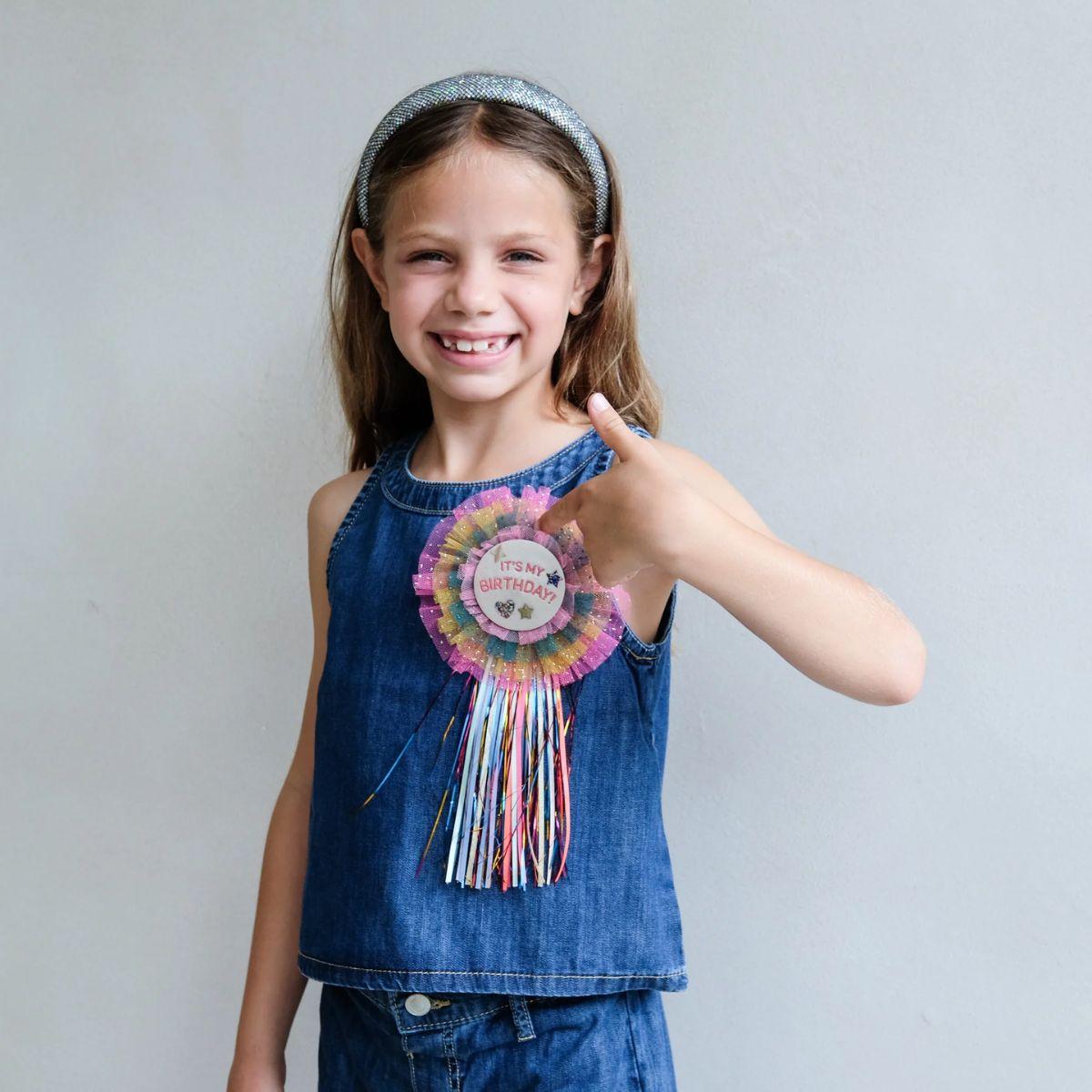 Young girl wearing a denim dress with a colorful birthday rosette, against a plain background