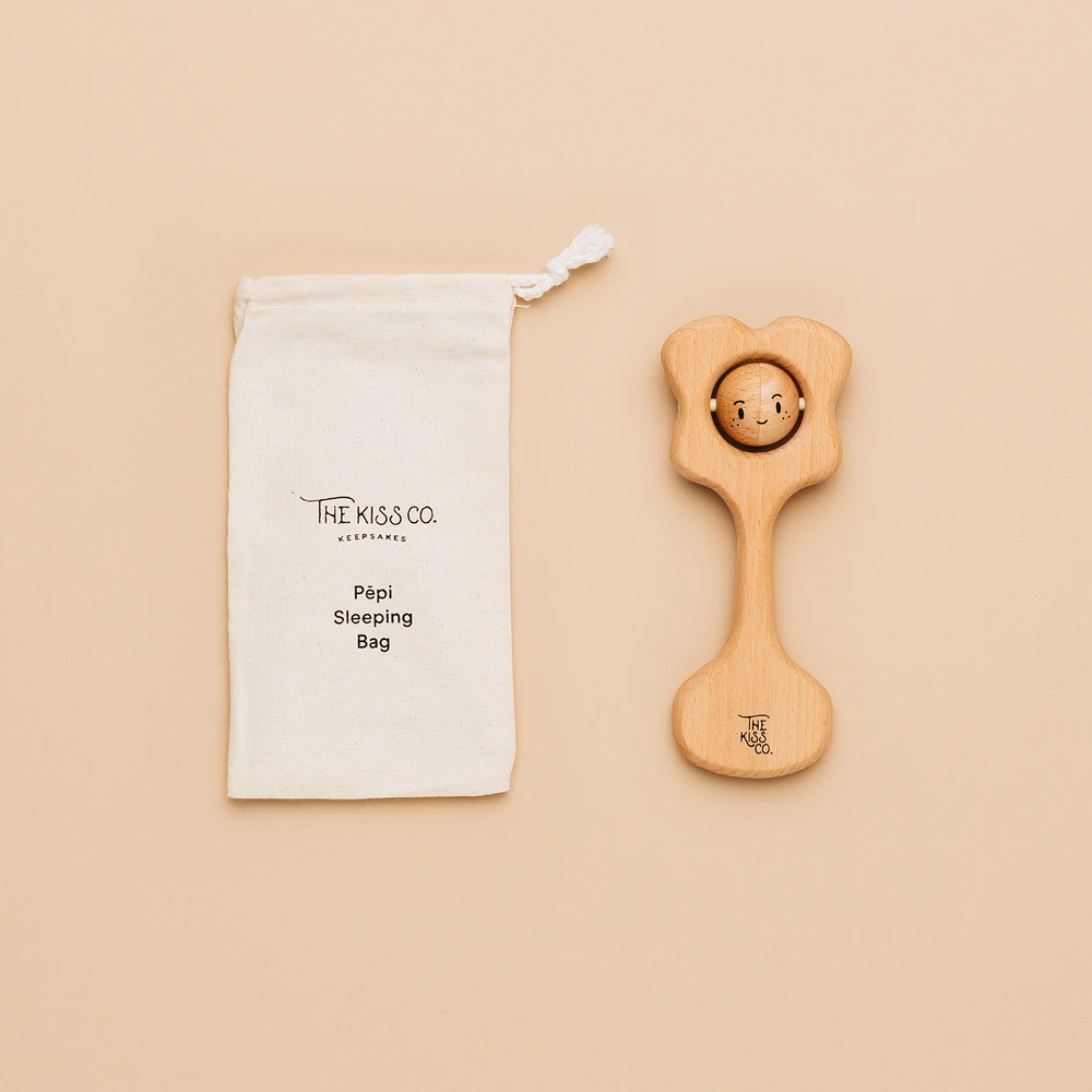 A natural beeswax-coated wooden teething rattle with a label attached reading 'Pǩpi Kiss Teething Rattle', alongside a cotton storage bag.