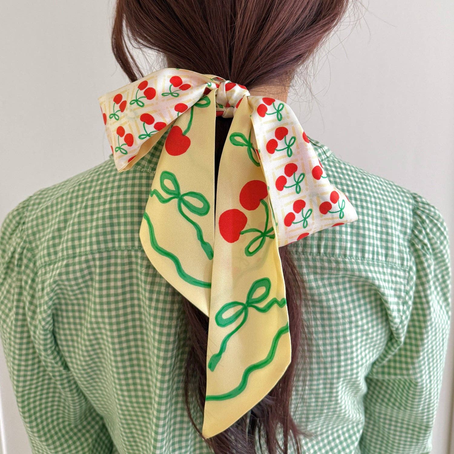 Person wearing a yellow hair scarf with red cherries and green bows, tied in a ponytail.