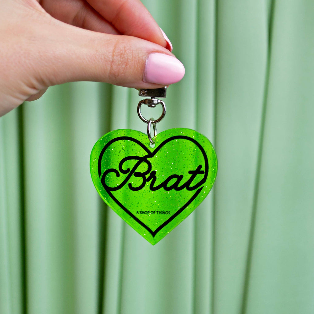 Heart-shaped keychain with 'Brat' text held by a hand against a green curtain background