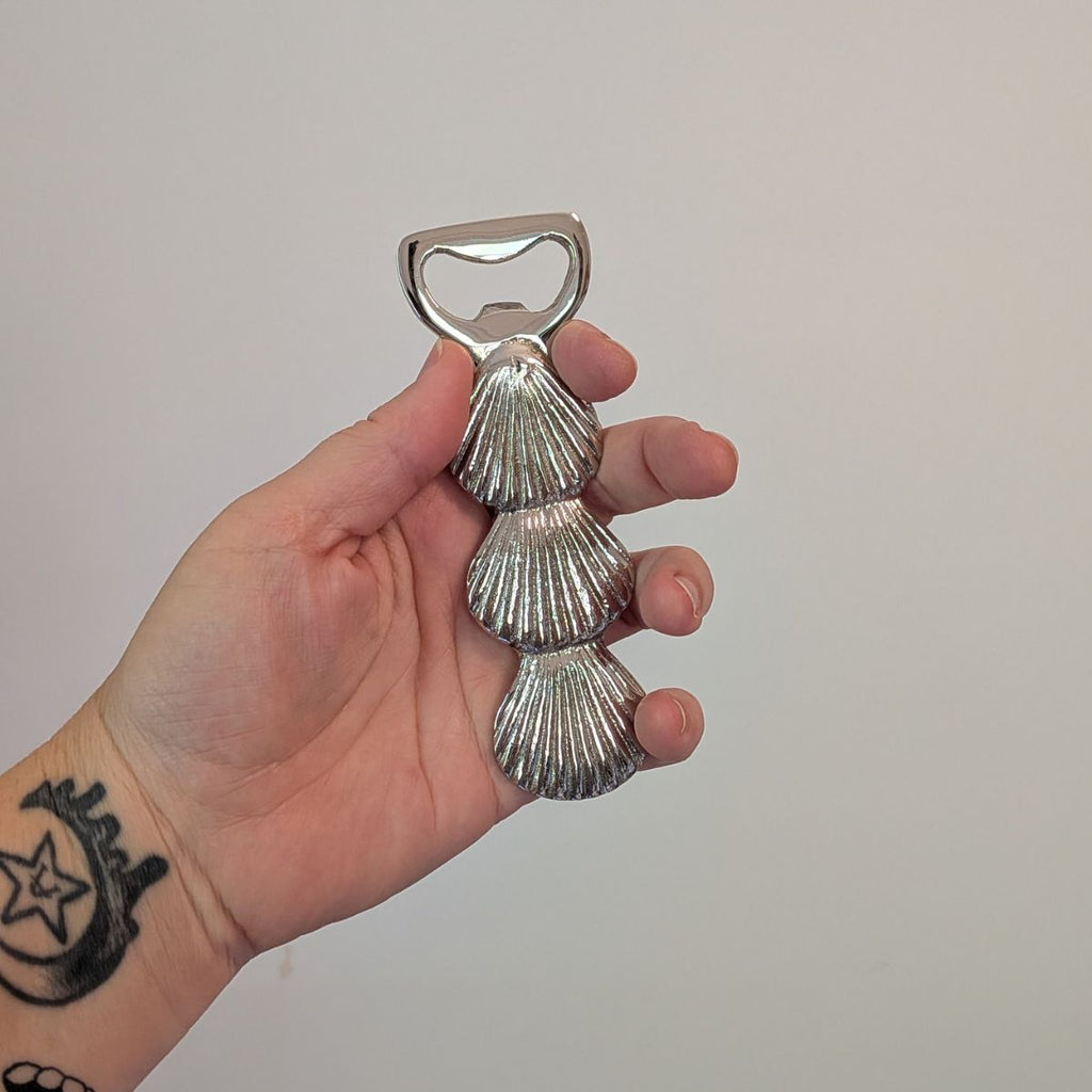 Hand holding a silver shell-shaped bottle opener against a plain background