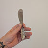 Hand holding a butter knife with a decorative feather handle against a plain background