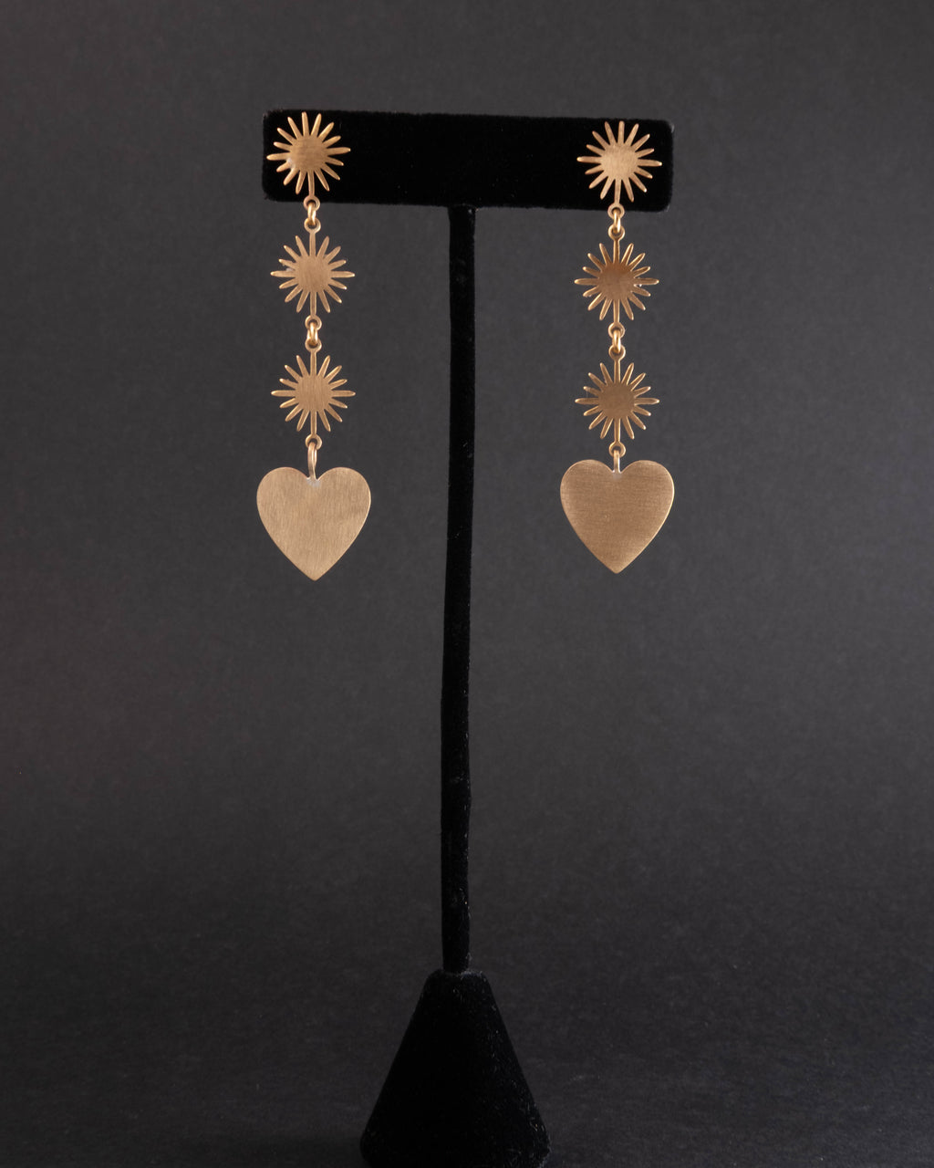 Brass Gold heart-shaped earrings with star-like top elements on a black stand against a dark background