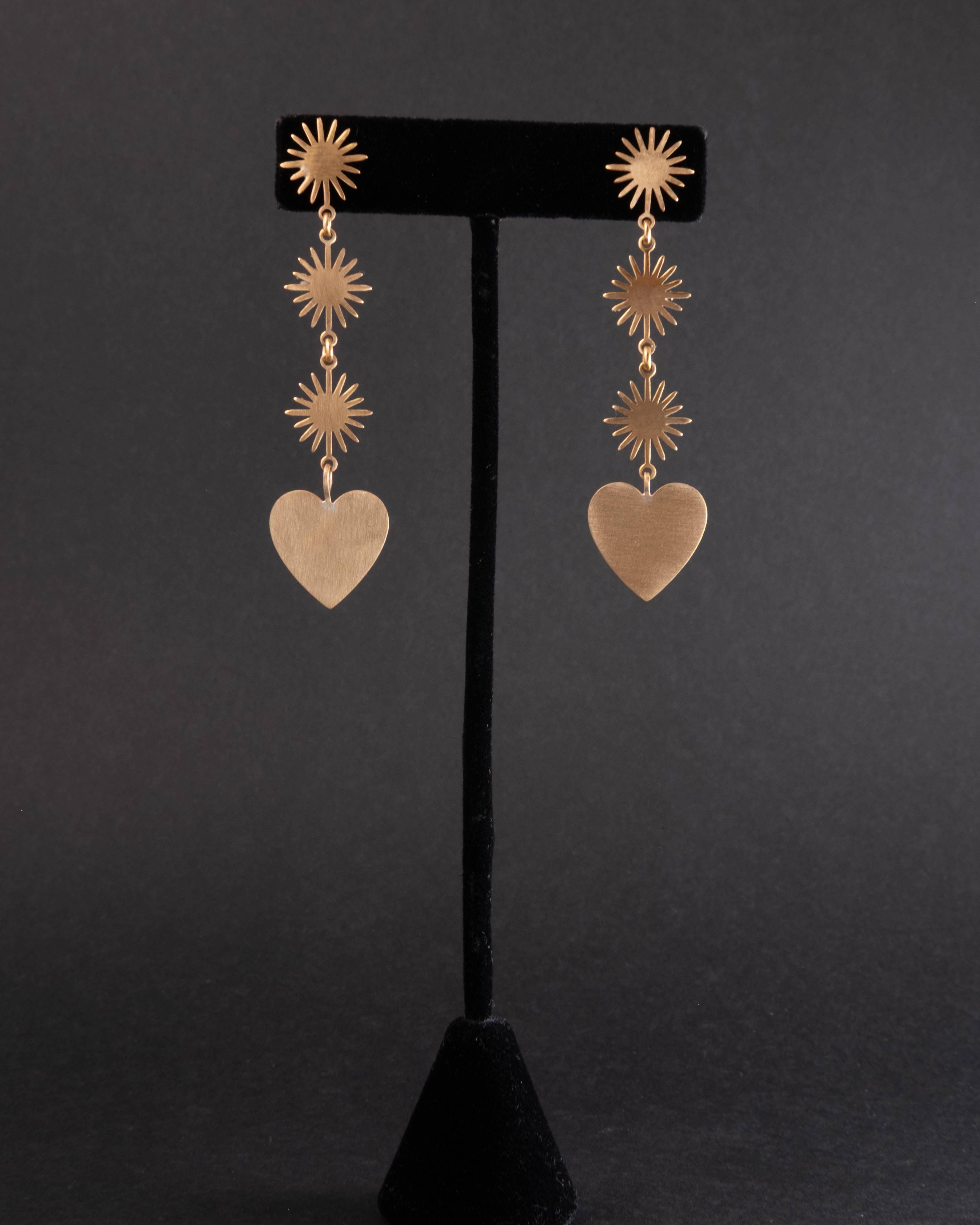 Brass Gold heart-shaped earrings with star-like top elements on a black stand against a dark background