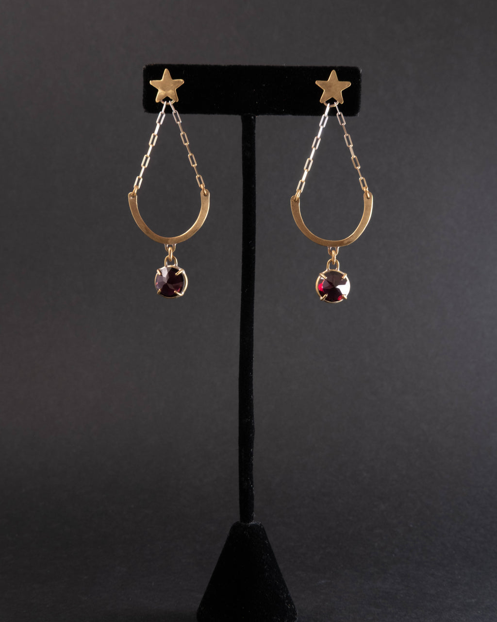 Pair of gold earrings with star and garnet gemstone design on a black stand against a dark background