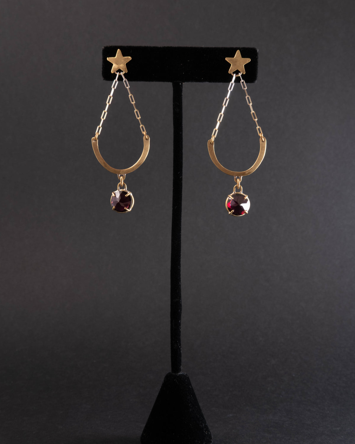 Pair of gold earrings with star and garnet gemstone design on a black stand against a dark background