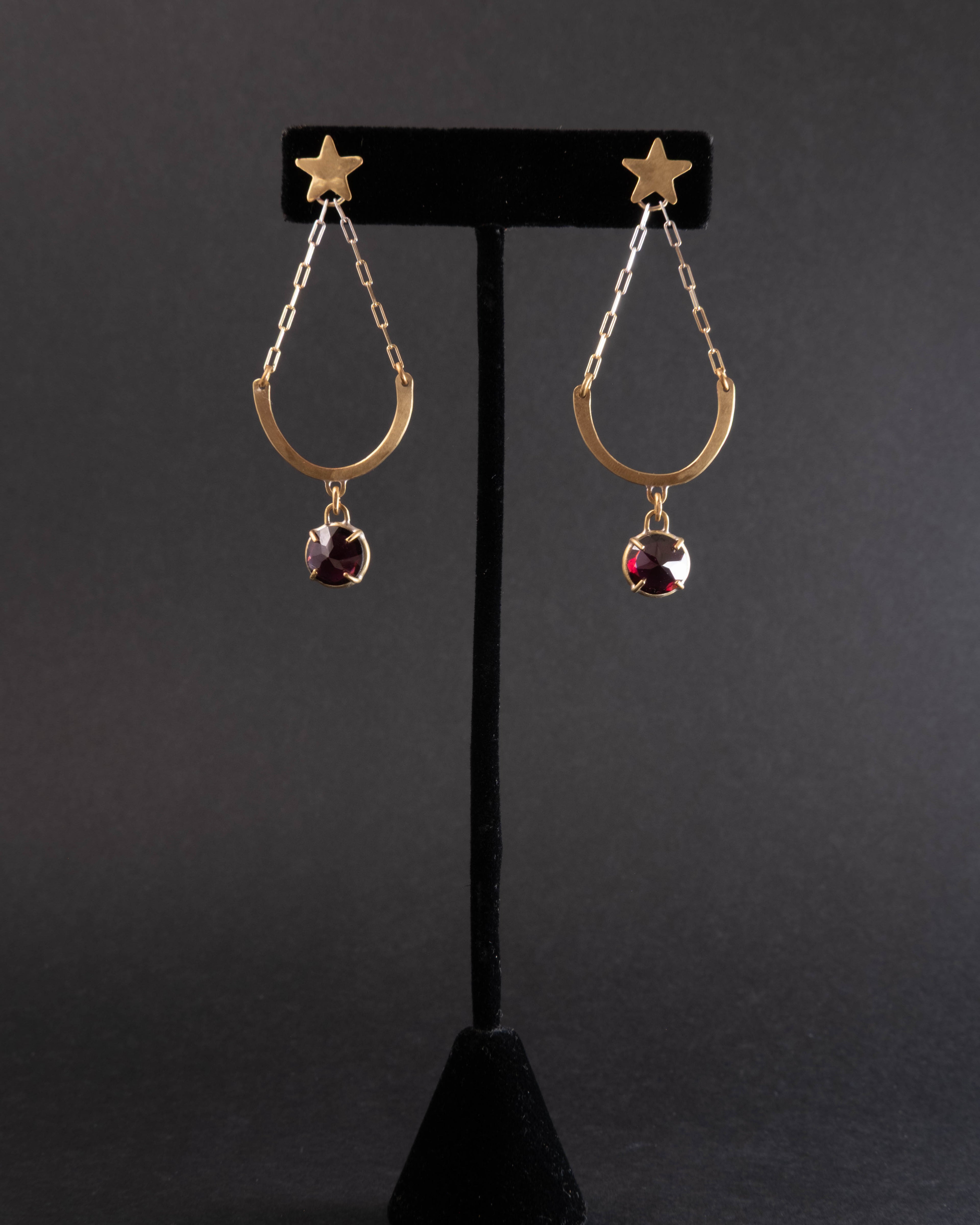Pair of gold earrings with star and garnet gemstone design on a black stand against a dark background