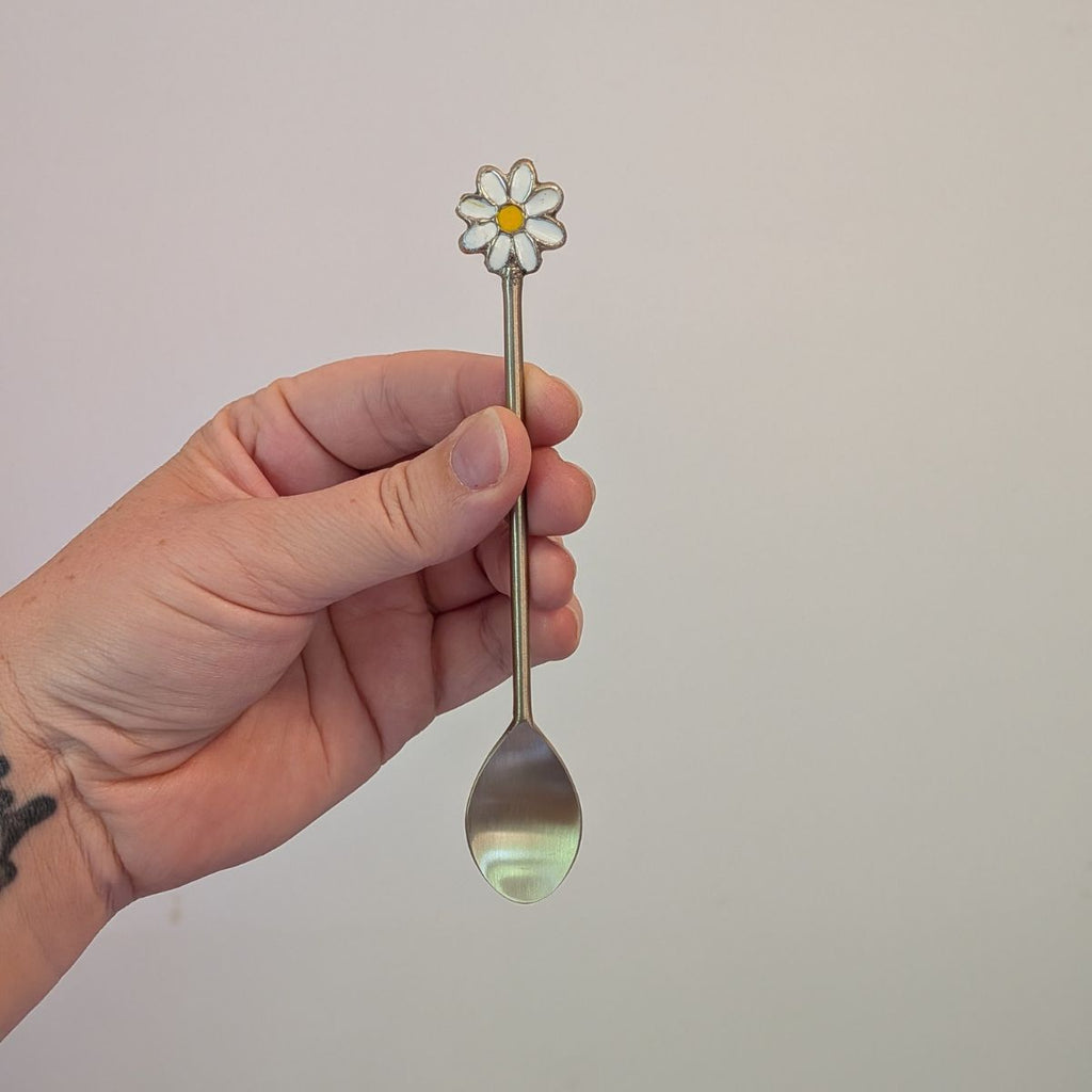 Hand holding a spoon with a daisy-shaped handle against a plain background