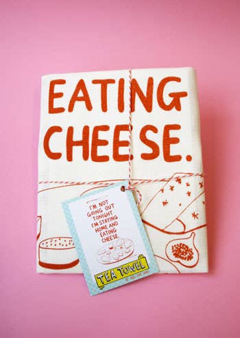 On a pink background, a folded cream-coloured tea towel with red writing and illustrations reads "Eating Cheese". 