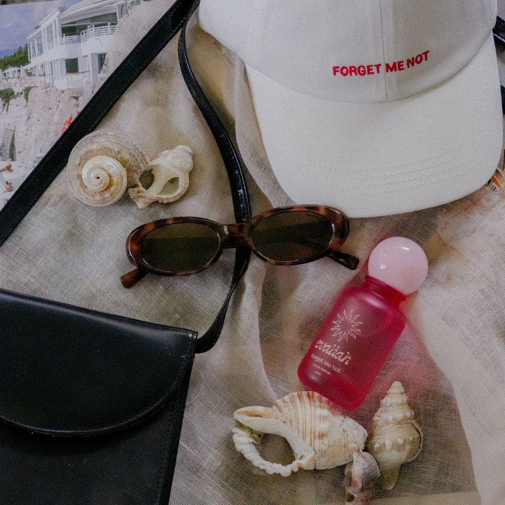 Beige cap with 'FORGET ME NOT' text, sunglasses, pink bottle, and seashells on a textured surface.