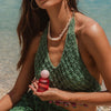 Woman in green crochet bikini top holding a red bottle with a white cap on a beach.