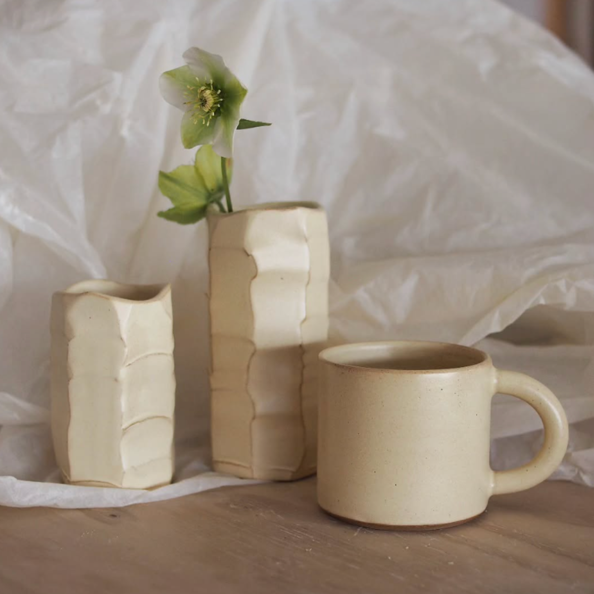 Three ceramic pieces including a mug and two vases on a wooden surface with a white fabric background.