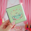 Compact mirror with a design of a fairy in a blue dress surrounded by flowers, held against a pink curtain background.